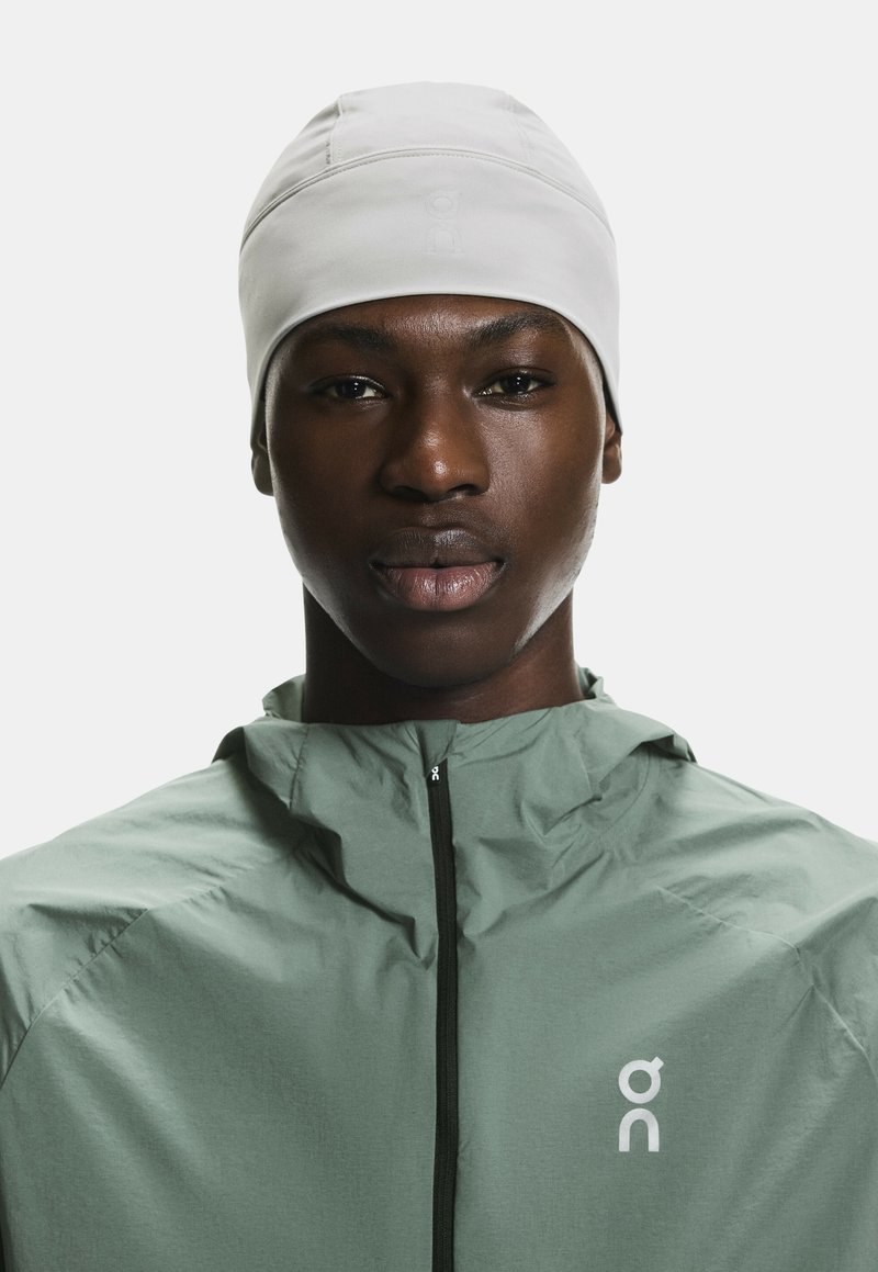 Jeune personne portant un bonnet blanc et une veste verte zippée avec le logo On sur la poitrine, face à un fond blanc.