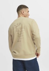 Beżowy sweatshirt z graficznym nadrukiem "Bucket List" z tyłu, zawierającym ilustracje miejsc i tekst. Żebrowane mankiety i dół.