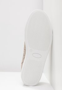 Semelle de sneaker blanche avec un motif de bande de roulement ondulé, marquée taille 37, positionnée verticalement contre un mur et un sol blancs.