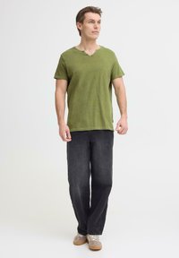 Luźna zielona oliwkowa koszulka w serek; zestawiona z czarnymi spodniami o prostych nogawkach oraz jasnymi sneakersami, wykonanymi z fakturowanej tkaniny.