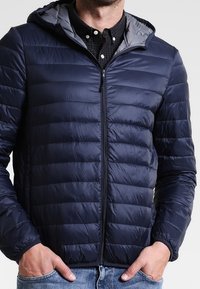 Marinblå pufferjacka med dragkedja fram, huva och grått foder. Har kviltad textur och elastiska ärmslut, bärs över en svart rutad skjorta.