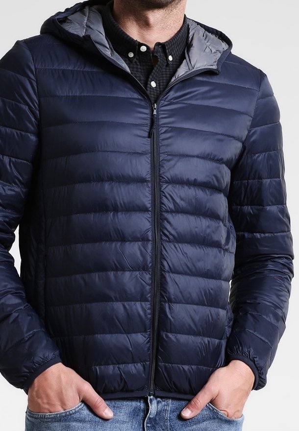 Marinblå pufferjacka med dragkedja fram, huva och grått foder. Har kviltad textur och elastiska ärmslut, bärs över en svart rutad skjorta.