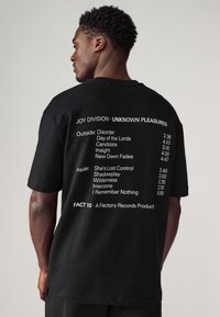 Black short-sleeve t-shirt featuring white printed text listing tracks from "Unknown Pleasures" by Joy Division, with a relaxed fit and casual design.
