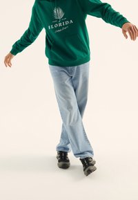 Sudadera verde con capucha y estampado blanco, jeans de pierna ancha azul claro, y zapatillas deportivas negras con detalles grises, mostradas en una pose dinámica.