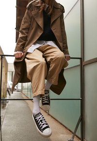 Trench-coat marron porté sur un pull marine, pantalon large couleur sable, et baskets noires aux lacets blancs. Fond gris et vert.