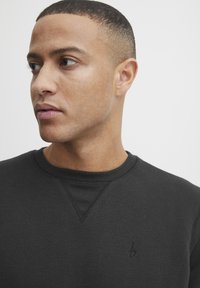 Sudadera negra con cuello acanalado y un panel de hombro texturizado. Logo bordado sutil en el pecho. Textura de tela suave.
