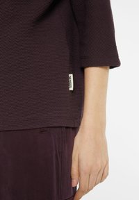 Lila strukturierte Bluse mit lockerer Passform, ausgestattet mit einem umgeschlagenen Ärmel und einem kleinen Etikett mit der Aufschrift "SENSES" an der Seite.