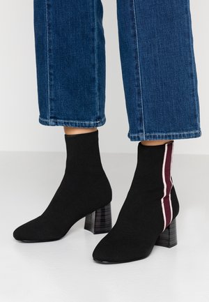 Schwarze gestrickte Stiefeletten mit Blockabsätzen und seitlichen Streifen in Weiß und Burgunder, getragen zu gekürzten blauen Jeans.