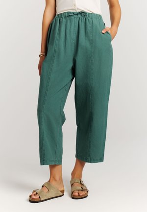 Vrouw draagt teal wijde cropped broek met elastische tailleband en beige slip-on sandalen met gespen.