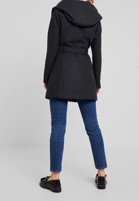 Cappotto nero di lana con ampio cappuccio, vita con cintura e tasche laterali con zip, abbinato a jeans di denim blu e mocassini neri.