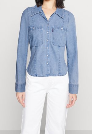 Persona che indossa una camicia di jeans blu aderente con bottoni anteriori e tasche con patta, abbinata a pantaloni bianchi, mostrata dal collo fino ai fianchi.