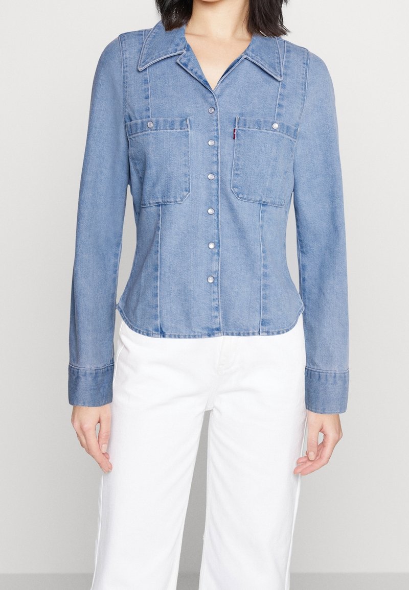 Personne portant une chemise en jean bleue ajustée avec des boutons sur le devant et des poches à rabat, associée à un pantalon blanc, vue du cou jusqu’aux hanches.