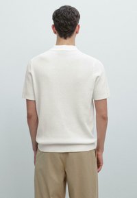 Polo bianca a maniche corte con un design a maglia testurizzata, caratterizzata da un colletto e un orlo leggermente a coste, indossata con pantaloni beige.