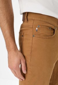Pantaloni in cotone marrone con vestibilità slim, tasche frontali e dettagli in metallo. La texture è liscia con una leggera lucentezza.