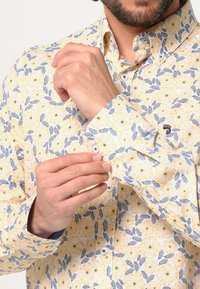 Korte mouw shirt met een geel bloemenpatroon op een witte achtergrond, met blauwe accenten en knoopsdetails op de manchetten en kraag.