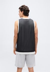 Νέος άνδρας που στέκεται με την πλάτη, φορώντας μαύρη αθλητική φανέλα χωρίς μανίκια από διαπνέον ύφασμα πάνω από λευκή φανέλα και ανοιχτό γκρι σορτς, μπροστά σε απλό φόντο.