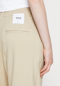 Beige broek van stof, met een achterzak en een wit label met de tekst "REPLAY ESSENTIAL". Soepele textuur en rechte snit.
