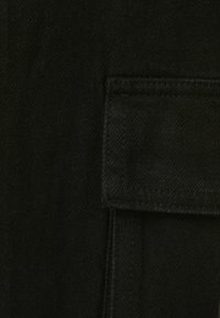 Tessuto denim nero con tasca a toppa, caratterizzato da una superficie liscia e dettagli di cucitura sottili. Lo sfondo è uniforme e scuro.