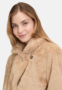 Cappotto in pelliccia sintetica beige con ampio colletto, dotato di abbottonatura e superficie morbida e strutturata.