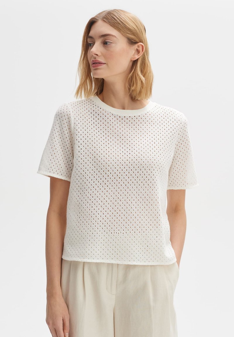 White knitted t-shirt with a crew neckline, featuring a textured, open mesh pattern and short sleeves. Paired with light-colored, loose-fitting pants.
