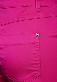 Fuksjowe spodnie o gładkiej fakturze, z tylną kieszenią i metalowym zatrzaskiem. Szwy są widoczne wzdłuż łączeń i krawędzi kieszeni.