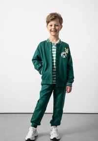 Zielona kurtka typu varsity z kołnierzem w paski, naszywką z logo, w połączeniu z dopasowanymi spodniami i polo w paski. Całość dopełniają białe trampki.