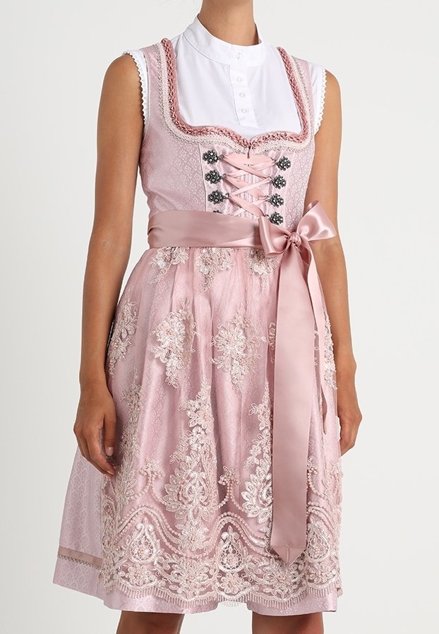 Robe traditionnelle rose avec superposition de dentelle fleurie, nœud en satin à la taille et blouse blanche boutonnée en dessous, portée par une personne debout.