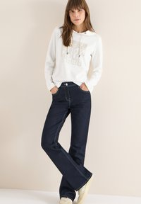 Mujer con una sudadera blanca de manga larga con texto, jeans de tiro alto de color azul oscuro, zapatillas beige, de pie contra un fondo beige liso.