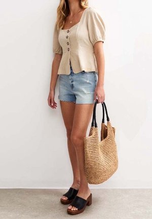 Femme portant un chemisier beige à manches bouffantes et un short en denim tient un grand sac cabas tressé, debout contre un mur blanc.