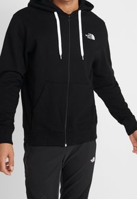 Zwarte zip-up hoodie van zachte stof, met een wit logo op de linkerkant van de borst, witte trekkoorden en voorzakken.