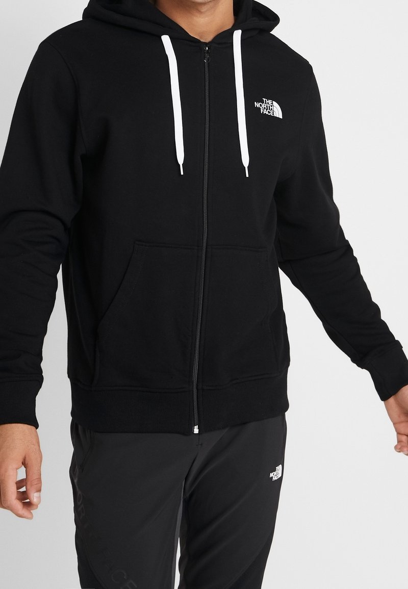 Zwarte zip-up hoodie van zachte stof, met een wit logo op de linkerkant van de borst, witte trekkoorden en voorzakken.