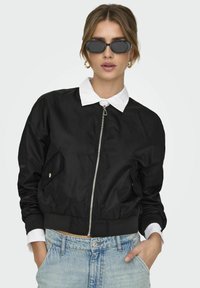 Veste bomber noire avec fermeture éclair, poches latérales, poignets et ourlet côtelés ; associée à un jean taille haute bleu clair et des lunettes de soleil.