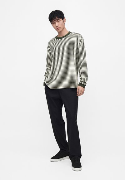 Jeune homme portant une chemise à manches longues rayée verte et blanche, pantalon noir et baskets noires, debout devant un fond blanc uni.