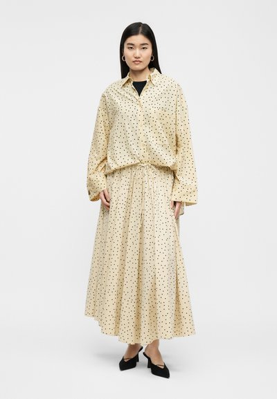 Femme portant une chemise beige à manches longues avec boutons et une jupe longue plissée assortie à pois noirs, talons noirs, debout dans une posture neutre.