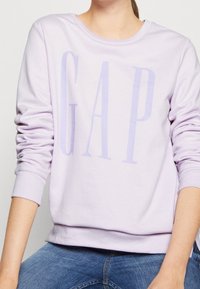 Felpa viola con grande logo "GAP", texture morbida, scollo rotondo e maniche lunghe con polsini, abbinata a jeans blu.
