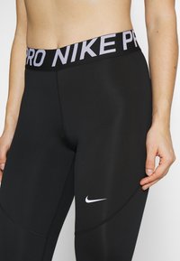 Mustat urheiluleggingsit, joissa on leveä vyötärönauha, jossa lukee "PRO NIKE" valkoisella, ja pieni valkoinen swoosh-logo nilkan lähellä. Sujuva pinta.