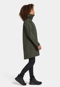 Eine lange olivgrüne wasserdichte Jacke mit hohem Kragen, seitlichen Taschen und geknöpften Manschetten, kombiniert mit schwarzen Leggings und schwarzen Stiefeln.