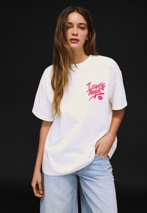 Young woman with long hair wearing a white oversized T-shirt with pink "Iconic Angel" text and light blue jeans against a black background.