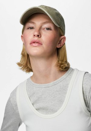 Jeune femme aux cheveux blonds courts porte une casquette beige, un t-shirt gris à manches longues et un haut blanc sans manches sur fond blanc.