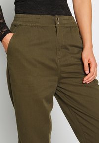 Olivové kalhoty s elastickým pasem, dvěma bočními kapsami a dvěma kovovými knoflíky vpředu. Hladká textilie.