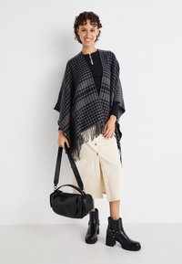 Schwarz-graues kariertes Poncho mit Fransen, getragen über einem schwarzen Oberteil, kombiniert mit einem cremefarbenen Rock und schwarzen, höheren Stiefeletten. Enthält eine schwarze Tasche.