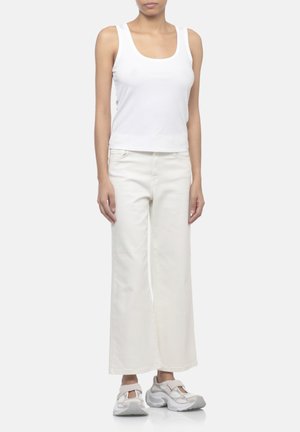 Donna che indossa un top bianco senza maniche, pantaloni bianchi a vita alta e gamba larga e sneakers bianche e grigie, in piedi davanti a uno sfondo uniforme.