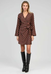 Femme portant une robe portefeuille rayée diagonale marron et noire avec de longues manches et des bottes noires jusqu'aux genoux, se tenant devant un fond uni.