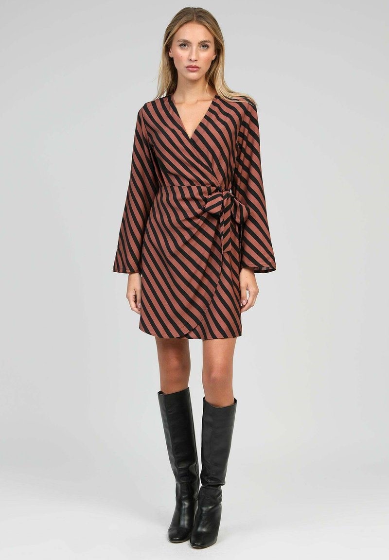 Femme portant une robe portefeuille rayée diagonale marron et noire avec de longues manches et des bottes noires jusqu'aux genoux, se tenant devant un fond uni.