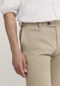 Beige Chinos aus strukturiertem Stoff mit flachem Vorderteil, Seitentaschen und Knopfverschluss am Bund. Kombiniert mit einem weißen Hemd.