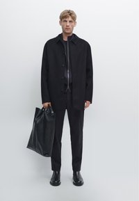 Cappotto di lana nera con colletto, indossato su un abbigliamento scuro, abbinato a stivali neri in pelle eleganti, mentre tiene una borsa a tracolla nera con texture.