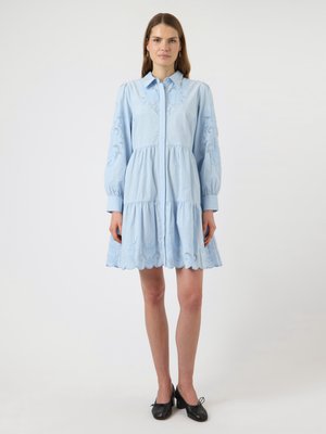 Kobieta w jasno niebieskiej sukience z długim rękawem, z haftowanymi kwiatowymi wzorami, zapięta z przodu na guziki, w czarnych butach na niskim obcasie, stojąca na białym tle.