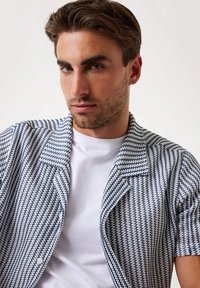Camisa de manga corta, con botones, con un patrón geométrico en azul y blanco. Tejido ligero, cuello con muesca y camiseta blanca contrastante debajo.