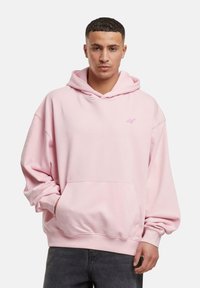 Sweat à capuche rose en matériau doux, avec une poche frontale, des poignets côtelés et un petit logo brodé sur la poitrine.