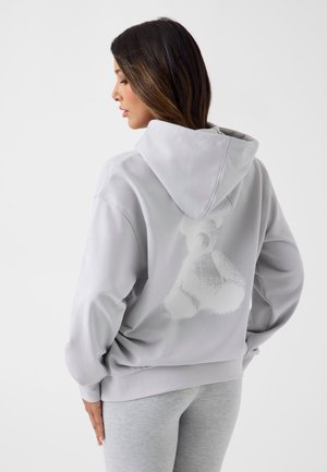 Donna con lunghi capelli scuri che indossa una felpa con cappuccio grigio chiaro con un tenue disegno di orsetto sulla schiena e leggings grigio chiaro abbinati.